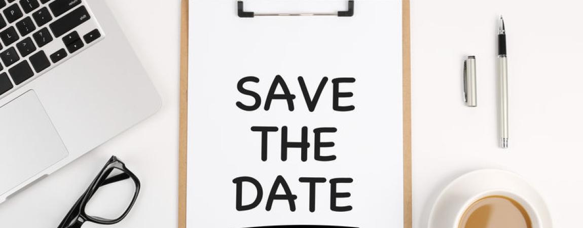 A clipboard with the words "Save The Date" written on a sheet of paper. Surrounding the clipboard on the desk are  aptop, glasses, pen, binder clip and cup of coffee.