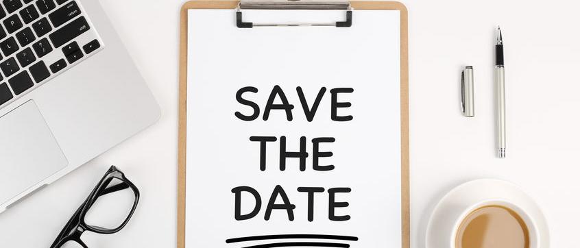 A clipboard with the words "Save The Date" written on a sheet of paper. Surrounding the clipboard on the desk are  aptop, glasses, pen, binder clip and cup of coffee.